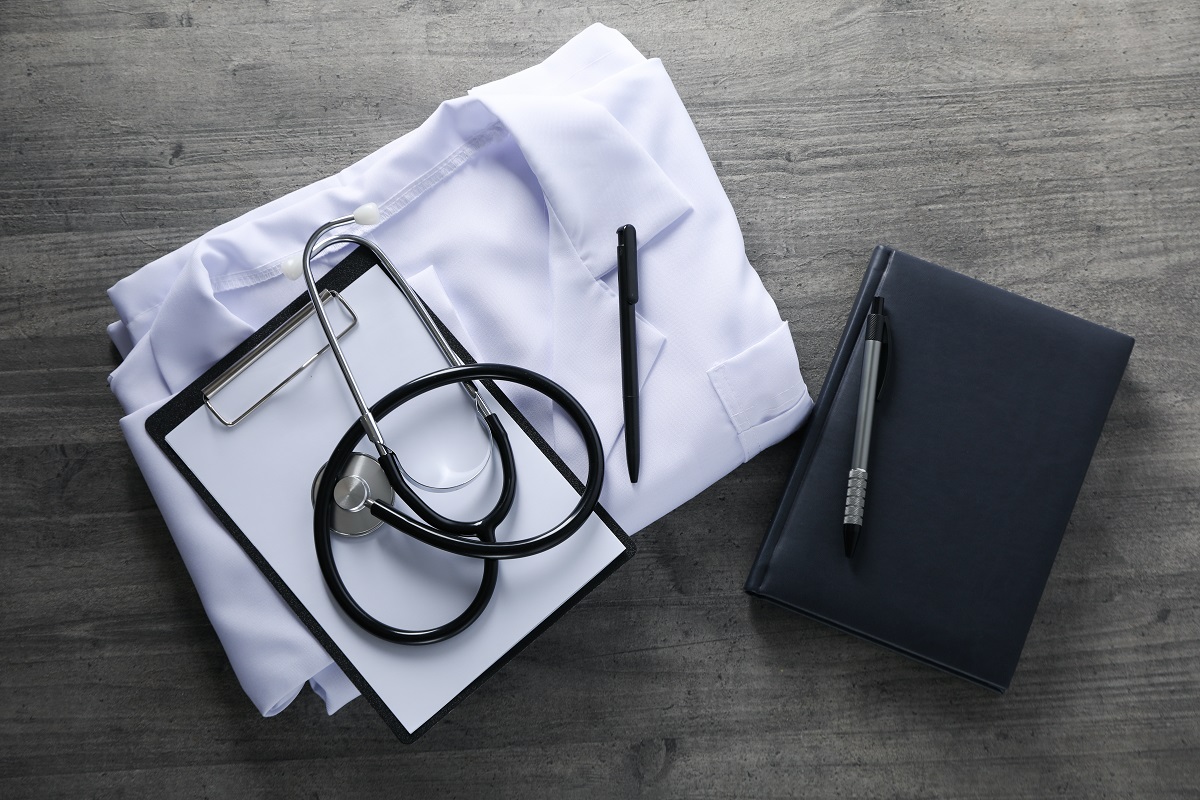 a folded doctor's coat, a stethoscope, medical notes, a notebook, and a pen on a wooden surface